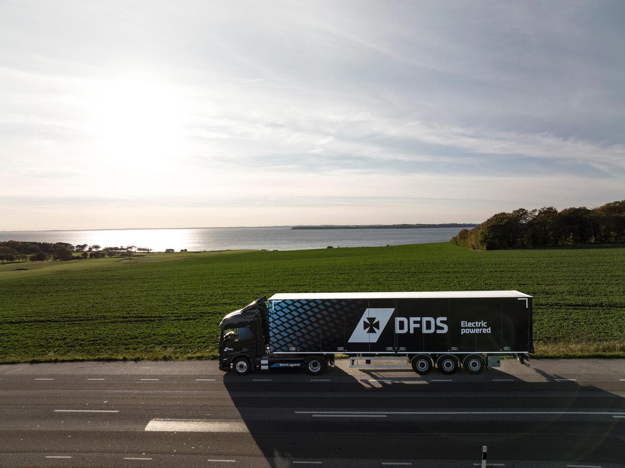 A drone picture of a dark blue truck with a trailer, seen from the left side, driving on the road in a green landscape, with water in the background