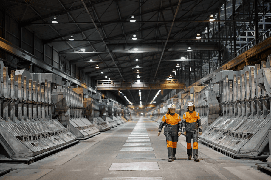 Två anställda i orangea arbetskläder går i en lagerlokal där metaller förvaras.